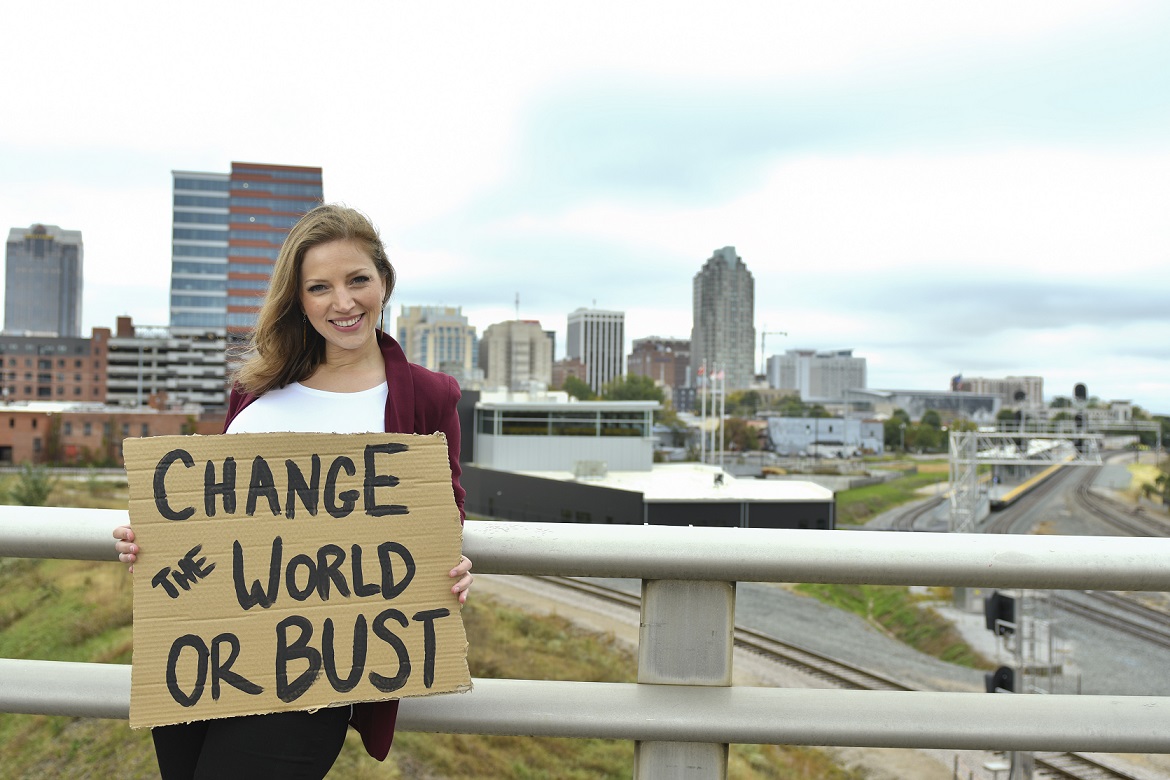 Amber Melanie Smith holding a sign that says Change the World or Bust against a city skyline